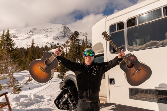 A musician holding two acoustic guitars outside next to a cat ski.